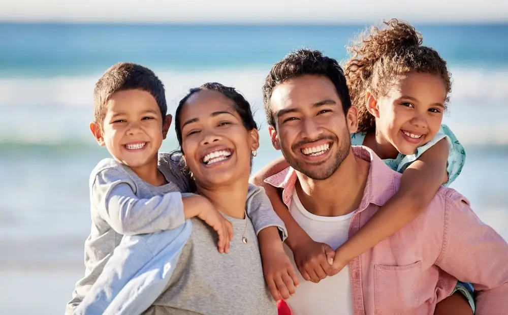 A family of four smiles at the camera on a sunny beach. The parents stand together while their two young children are playfully held on their backs. The sky is clear and the ocean waves are gently crashing in the background.