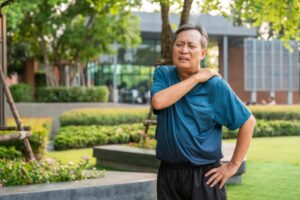 Middle-aged man in athletic wear standing outside in a park, holding his left shoulder and wincing in pain, suggesting discomfort or injury, with trees and a building in the background.