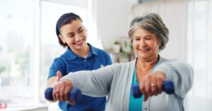 An older woman smiles while lifting small dumbbells as a younger female trainer in a blue shirt assists and encourages her in a bright, indoor setting.