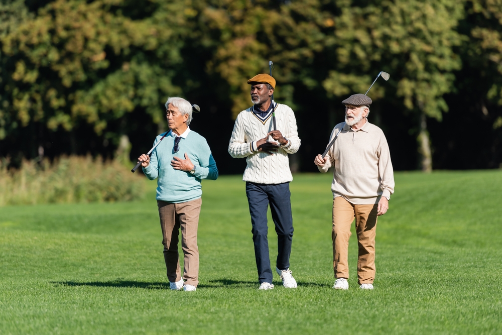 Three older adults, two men and one woman, walk across a golf course carrying golf clubs on their shoulders, dressed in casual golf attire, with trees and green grass in the background.