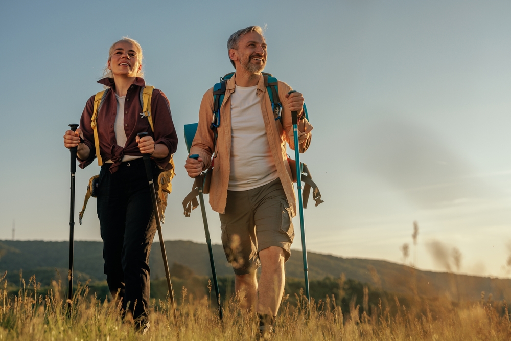 Two older adults with backpacks and hiking poles walk through a grassy field at sunset, smiling and enjoying the outdoors. Rolling hills and a clear sky are visible in the background.