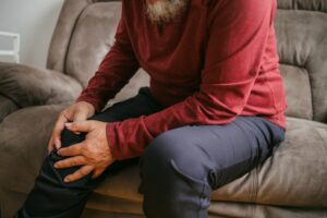An older adult wearing a red long-sleeve shirt and dark pants sits on a couch, holding their knee with both hands, appearing to be in pain or discomfort.