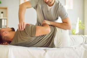A man lies on his side on a treatment table while a physical therapist in scrubs assists by gently stretching and adjusting his back and shoulder in a bright, comfortable clinic.